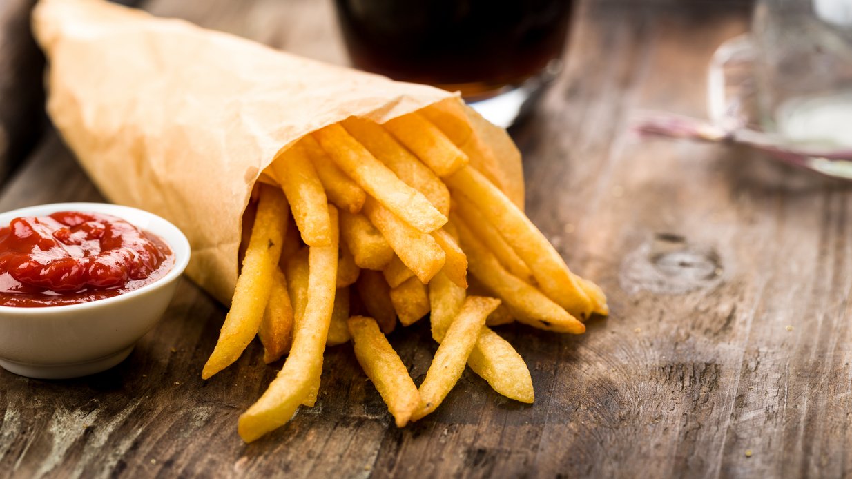 Papiertüte mit Pommes und einem Schälchen Ketchup auf einer Holztischplatte