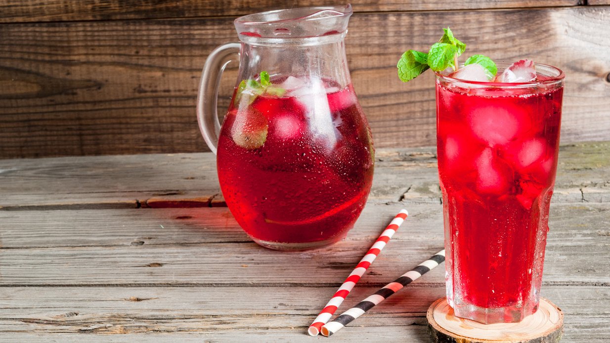 Rotes Getränk in Glas und -kanne mit Eiswürfeln und Minze garniert. Dazwischen zwei geringelte Pappstrohhalme, alles auf einem Holztisch drapiert.